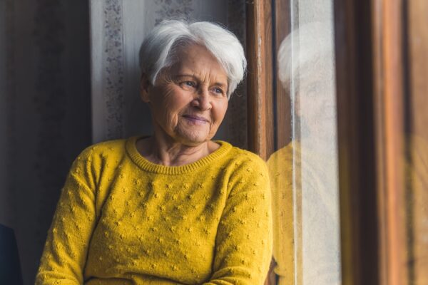 A senior woman in a yellow sweater looks out her window