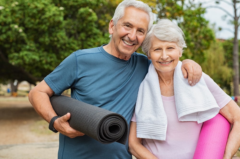 A senior couple smiles together while holding their yoga mats