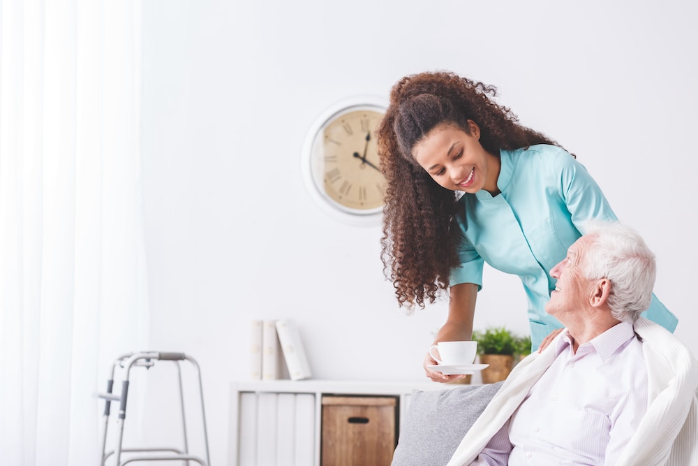 A happy young caregiver brings a senior man a cup of tea