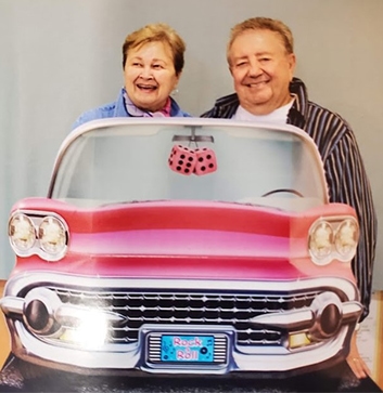 Marilyn and David Crossley share a moment posing for a photograph behind an image of a 1950s-style automobile.