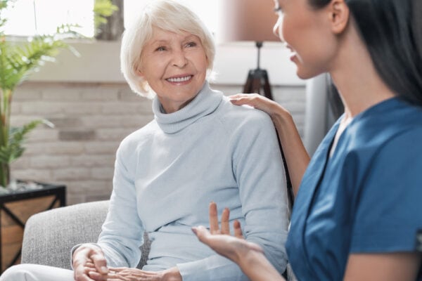 A senior woman and a female respite care nurse sit and talk on a sofa