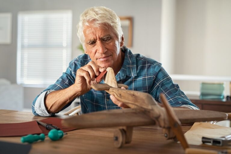 Focused,Senior,Man,Using,Sandpaper,To,Polish,Wooden,Airplane,Sculpture.