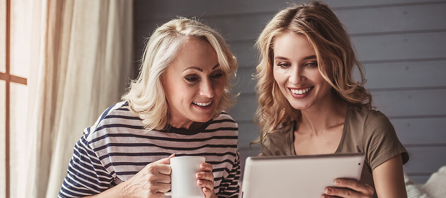 Beautiful senior mom and her adult daughter are using a digital tablet and smiling while sitting on couch at home