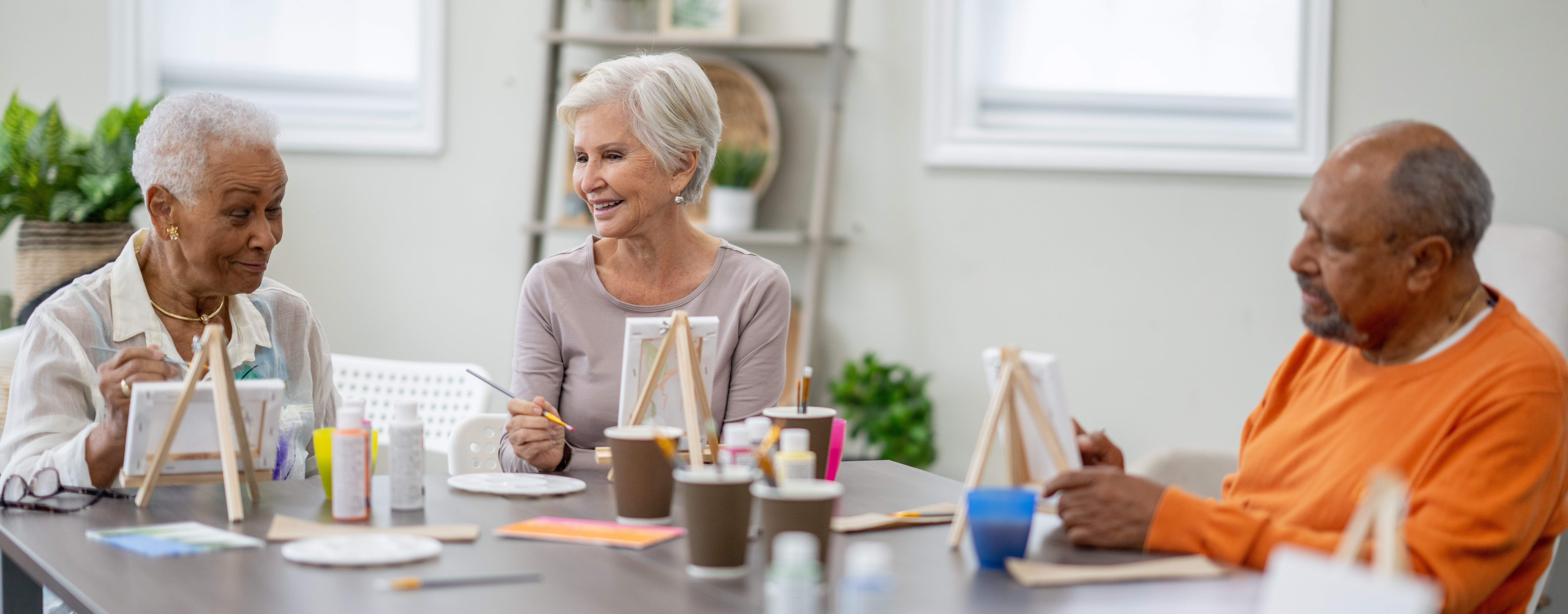 A small group of seniors are seen sitting around a large table as they paint together during an activity time in a retirement residence. They are each dressed casually and appear to be having fun.