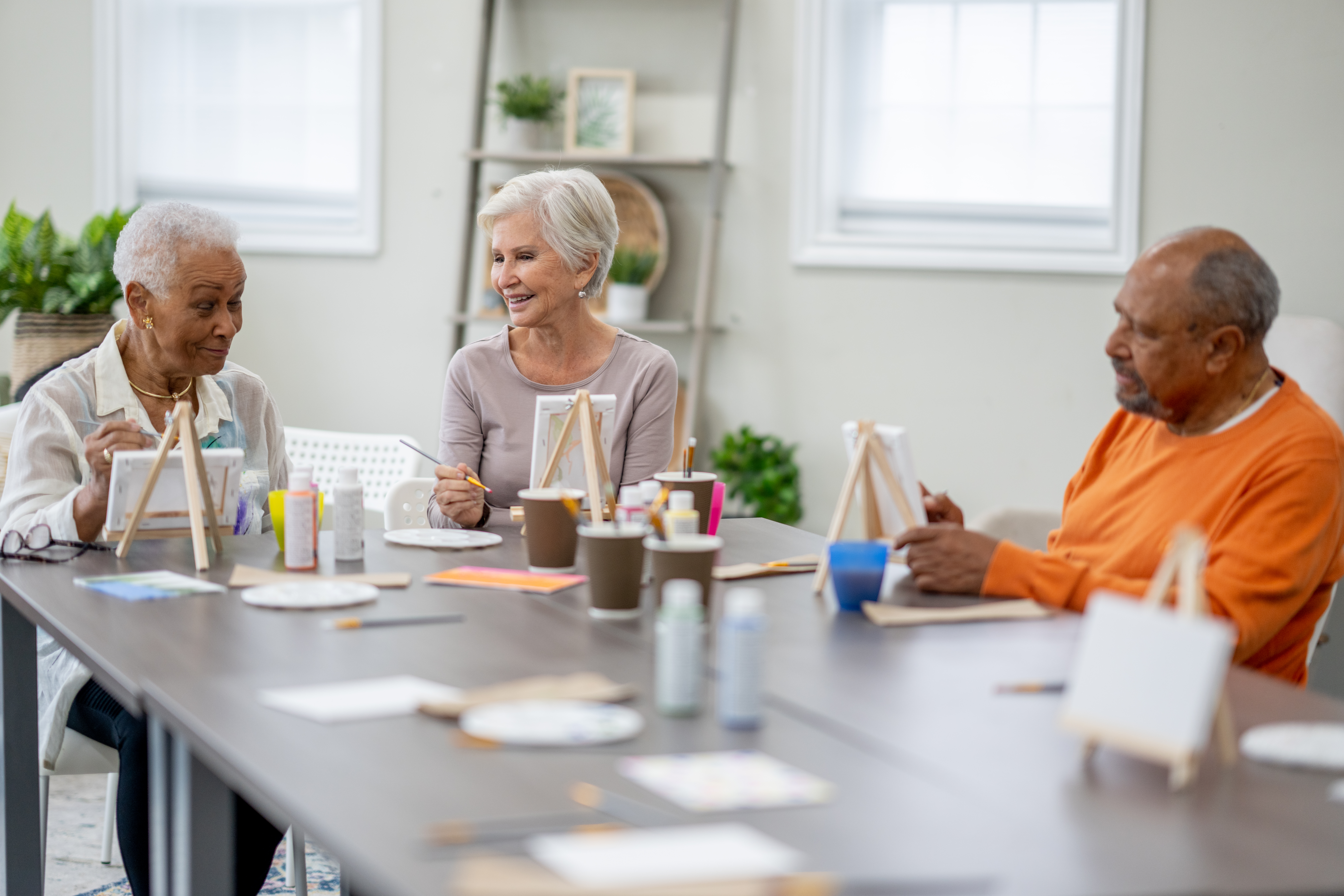 A small group of seniors are seen sitting around a large table as they paint together during an activity time in a retirement residence. They are each dressed casually and appear to be having fun.