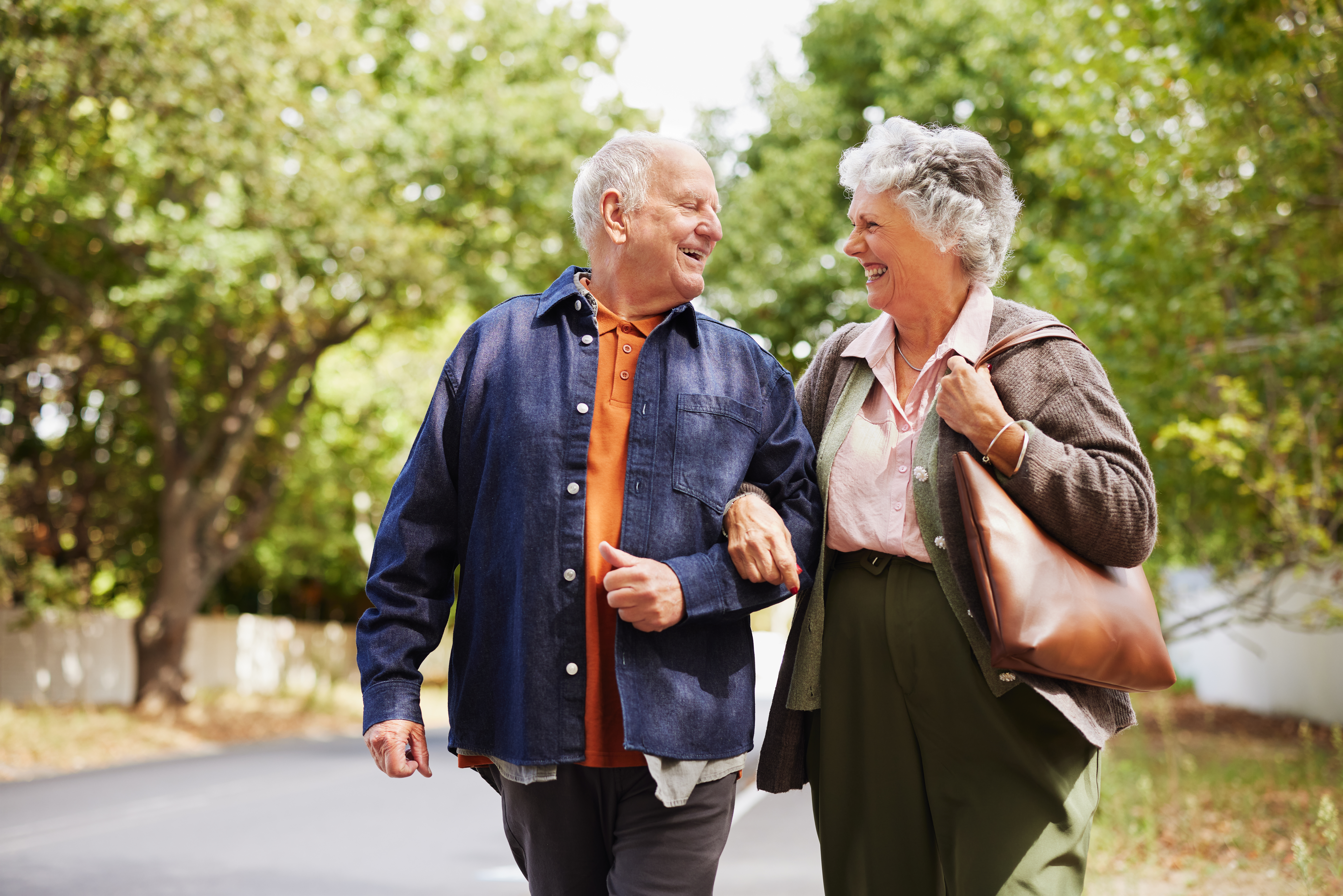 Smiling senior couple strolling together outside in a natural park. Joyful old man and laughing woman walking arm in arm on a sunny day with copy space. Happy elder couple enjoying a peaceful walk while looking at each other.