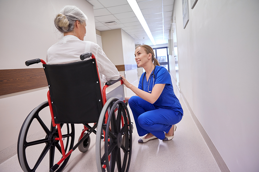 medicine, age, support, health care and people concept - happy nurse talking to senior woman patient in wheelchair at hospital corridor