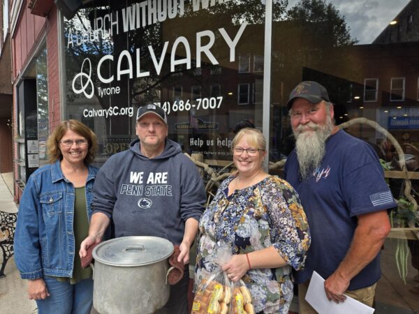Four adult volunteers, including staff from Colonial Courtyard at Tyrone, stand smiling in front of Calvary Church, holding a large metal pot of prepared food and a bag of bread rolls for the community meal outreach.