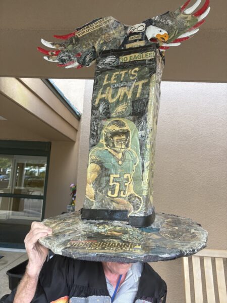 A person holding an extremely tall, elaborately decorated hat dedicated to the Philadelphia Eagles. The hat includes a large eagle sculpture on top, NFL logos, and a player photo on the central pillar.