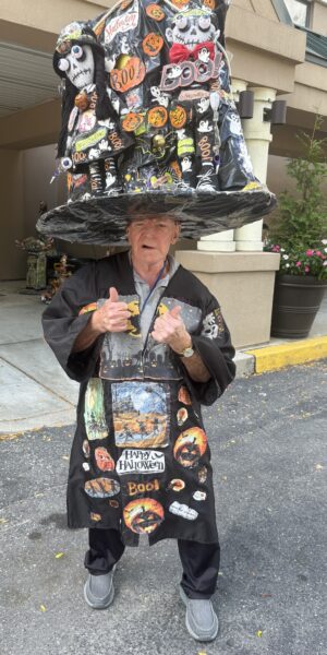 Independent Living resident Tom Papiernik, "The Hat Man," wearing an elaborate, oversized, handcrafted Halloween witch hat and matching tunic, giving two thumbs up.