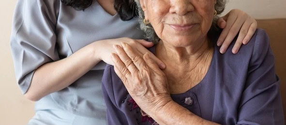 A caregiver, wearing a uniform, places a hand reassuringly on the shoulder and arm of a smiling elderly female resident.