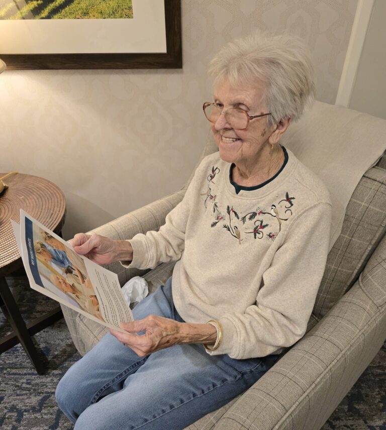 An elderly female resident at The Residence at Fitz Farm sitting in an armchair, smiling warmly while reading the community's Weekly Update newsletter.