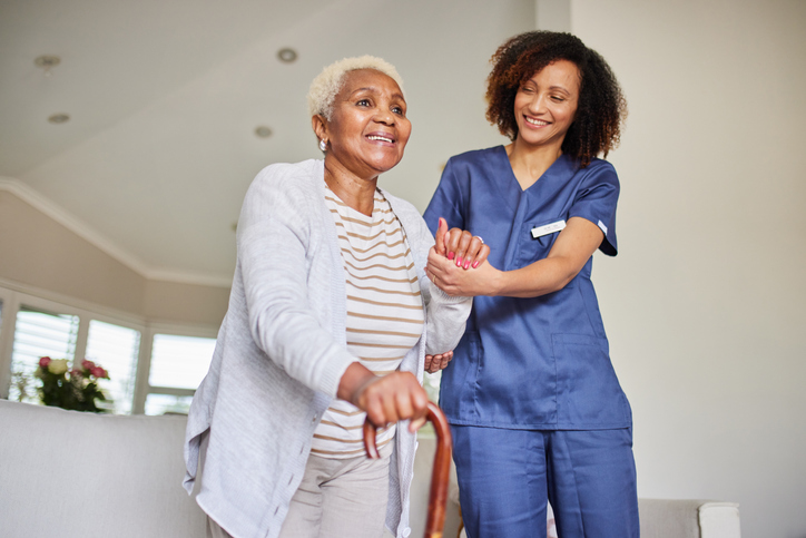 Warm scene of a caregiver assisting an elderly woman with a cane. Both are smiling, creating a heartwarming and supportive atmosphere indoors.