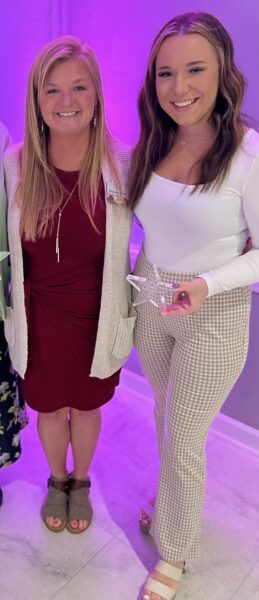 Clare Twentier (right) holds a star-shaped award while standing next to Krystle Pry, Executive Operations Officer at Newhaven Court at Clearview (left). Clare is wearing a white long-sleeved top and tan/white plaid pants, and Krystle is wearing a deep red dress with a light beige cardigan.