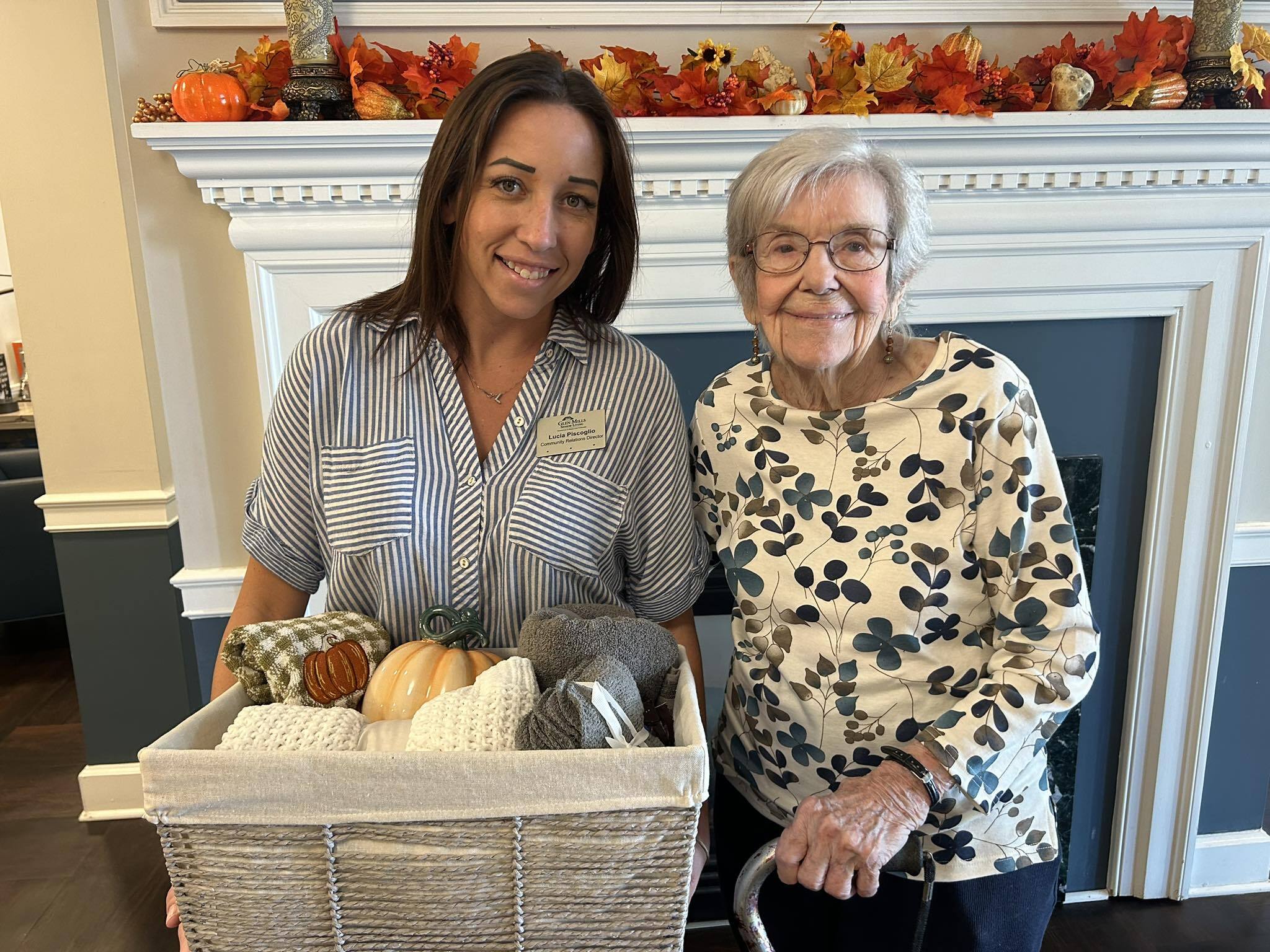 Lucia Piscoglio, Community Relations Director (CRD) at Glen Mills Senior Living, stands next to a resident, holding a basket filled with knitted items and fall decorations like pumpkins and autumn leaves.