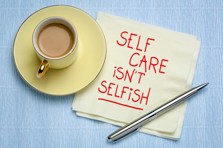 A top-down photograph showing a light yellow cup of coffee on a matching saucer next to a square, cream-colored napkin. Written on the napkin in red, handwritten-style capital letters is the phrase: SELF CARE ISN'T SELFISH. The phrase is underlined in red. A silver pen lies diagonally across the bottom-right corner of the napkin, and the items rest on a light-blue, textured surface.