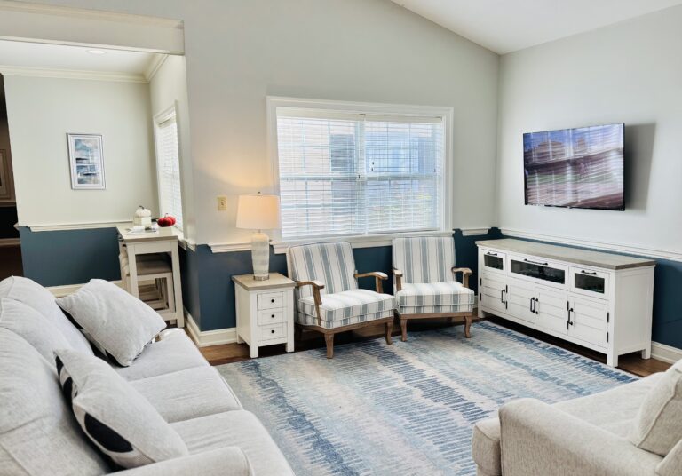 A sunlit living room in the lifebridges care neighborhood at Candle Light Cove, featuring coastal-style striped armchairs, a soft blue area rug, and a wall-mounted TV.