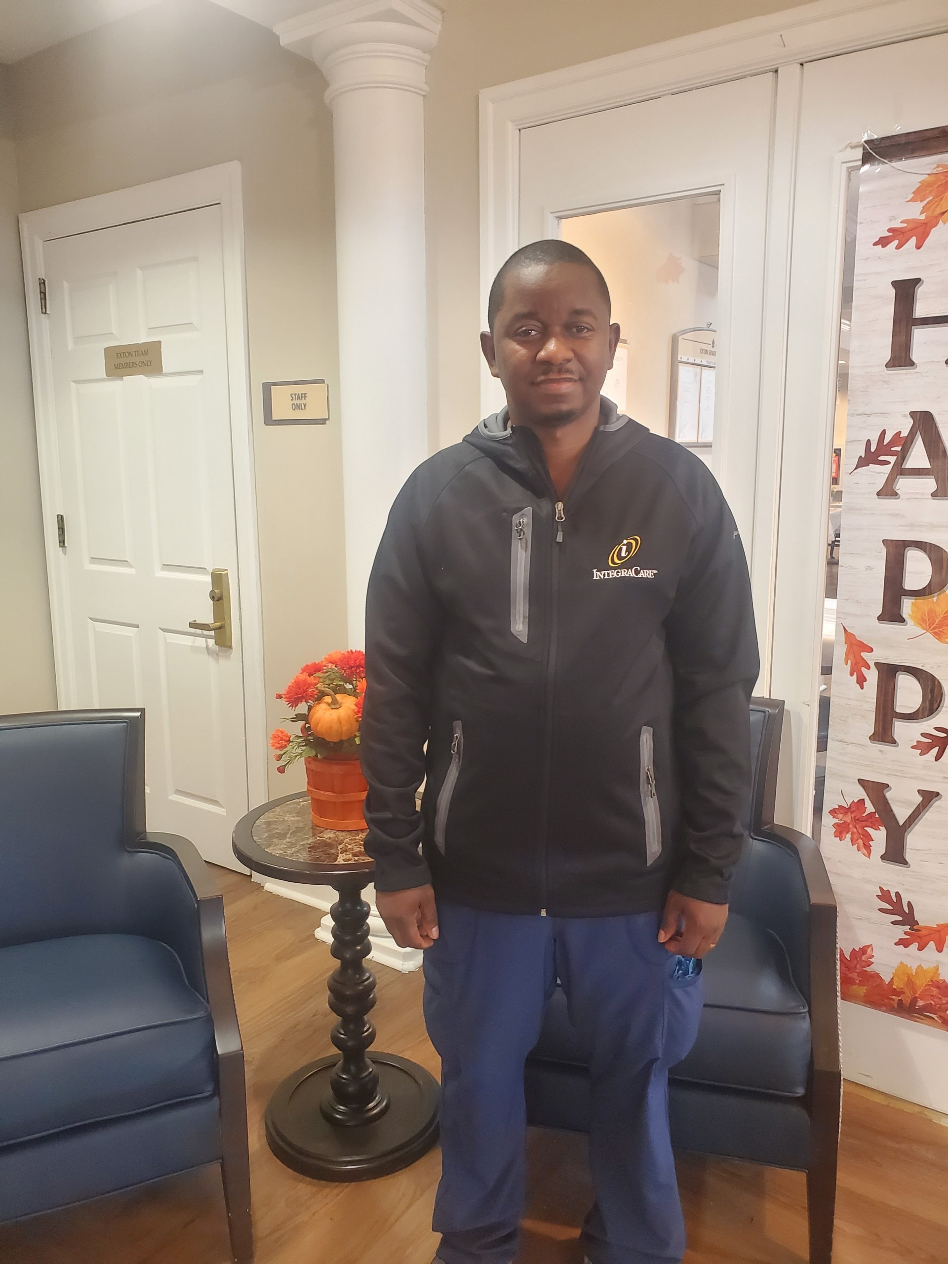 Mohamed Kargbo standing in the lobby of Exton Senior Living wearing a black IntegraCare jacket and blue scrubs, celebrating his 10-year anniversary.