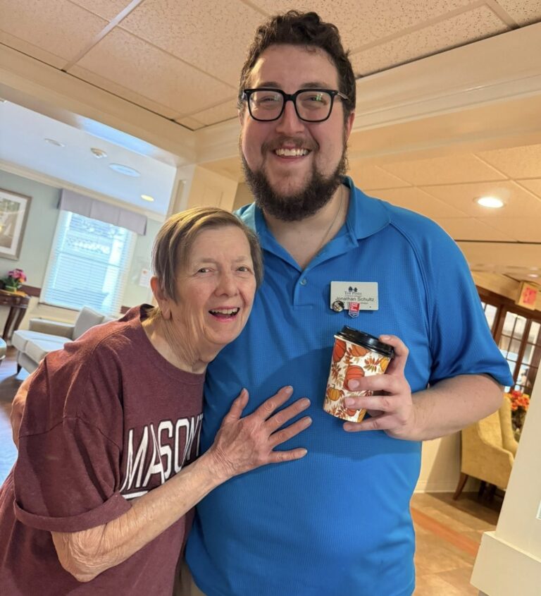 A smiling man with glasses, a beard, and a blue polo shirt standing next to an elderly woman in a maroon "MASON" t-shirt. The man, Jonathan Schultz, is holding a decorative coffee cup, and both are posing happily in a brightly lit indoor common area.