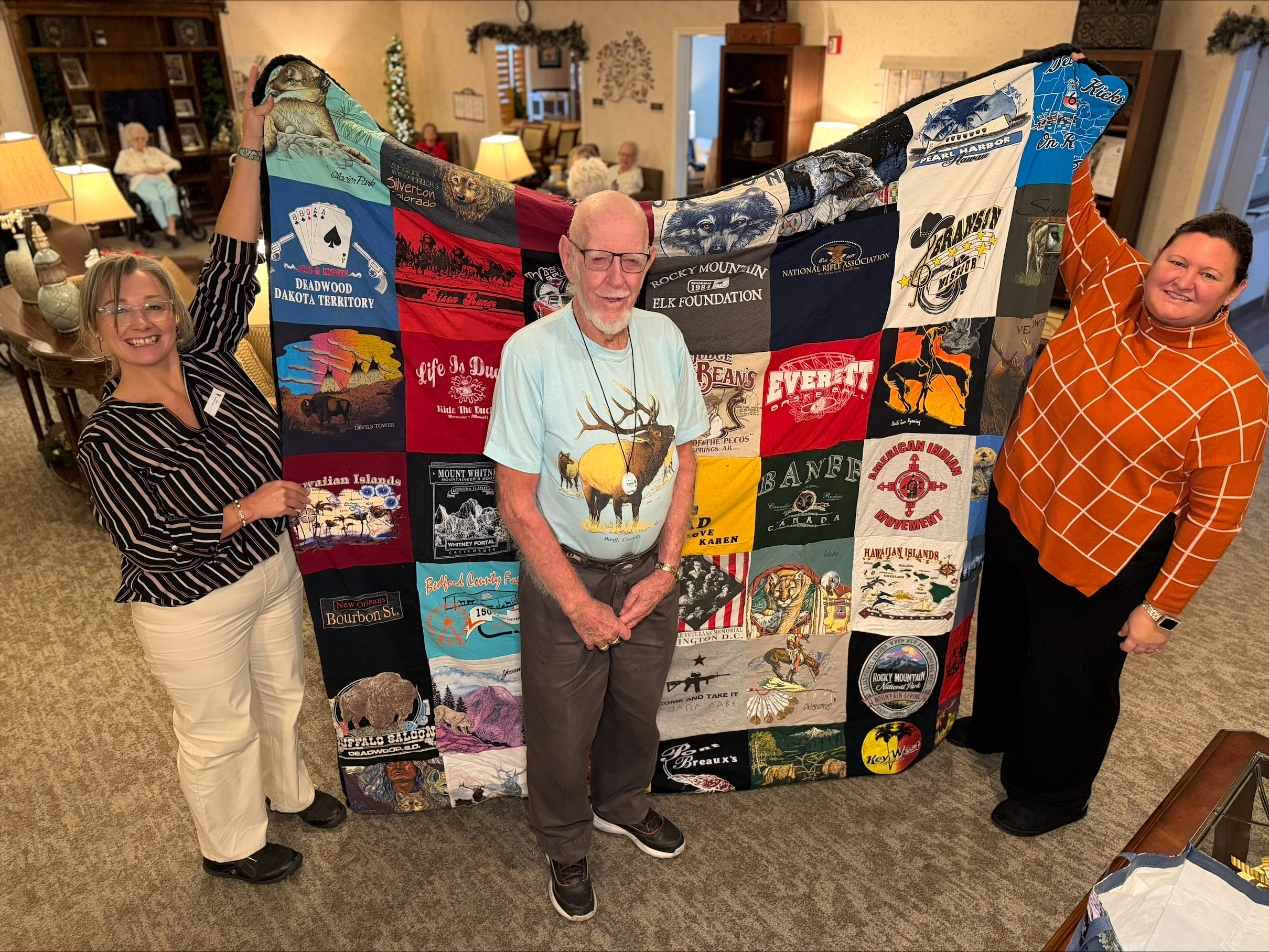 A man and two women showcasing a custom-made t-shirt memory quilt featuring travel and nature-themed graphics.